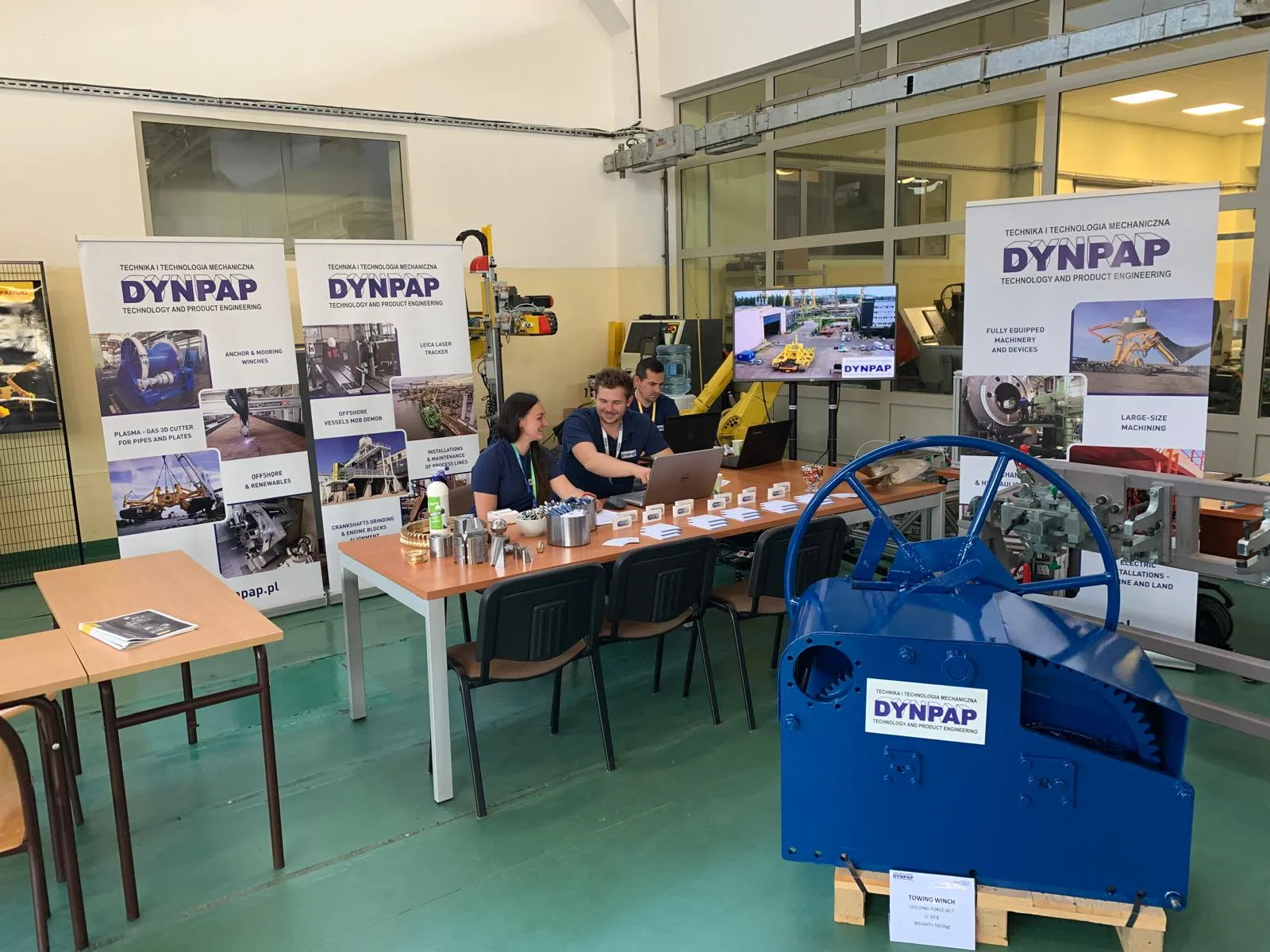 Stanowisko na targach z wieloma banerami DYNPAP. Przy stole siedzi trzech pracowników, jedna kobieta i dwóch mężczyzn. Uzgadniają coś przy komputerze.